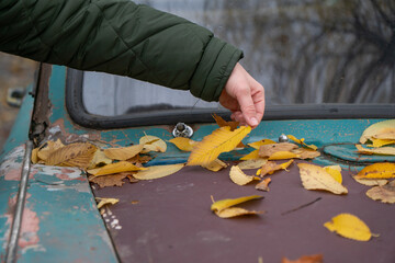 Hand gently touches yellow autumn leaves scattered on hood of weathered vintage car, showing peeling paint, muted green, brown tones, fall atmosphere