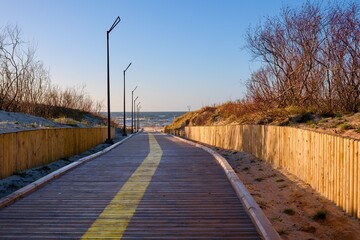 Wooden boardwalk leading through coastal dunes toward the sea. Empty seaside pathway with modern street lamps, perspective lines, and calm evening light, symbolizing travel, freedom, and nature.