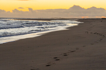 Footprints lead along the wet sand as waves roll in under the glow of a rising sun.