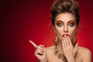 Glamorous woman surprised with colorful makeup and jewelry on red background  