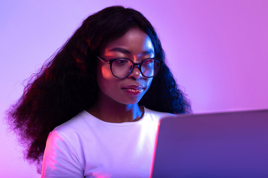 Focused millennial African American woman in eyeglasses engages with her laptop. She is studying remotely in a vibrant neon-lit environment, likely attending an online class or webinar. - Powered by Adobe