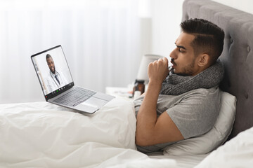 A sick Arab man sits in bed, wearing a scarf and using a laptop for a video call with a doctor. He is seeking medical advice from home during his illness, highlighting telemedicine's convenience.