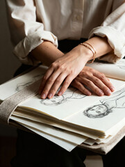 Obraz premium Artist's hands resting on a sketchbook with pencil portrait drawings. Close-up of a creative woman with her art portfolio. Hobby and passion concept