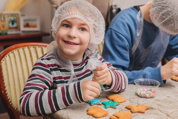 Portrait of a happy young boy in a hairnet and apron decorating gingerbread cookies with turquoise icing, joyful child participating in a festive Christmas baking workshop or culinary masterclass