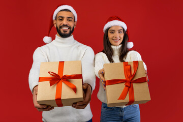 Fototapeta premium An arab couple, wearing Christmas hats, smiles joyfully while extending beautifully wrapped gifts towards the camera. The vibrant red background enhances their celebration of the winter holidays.
