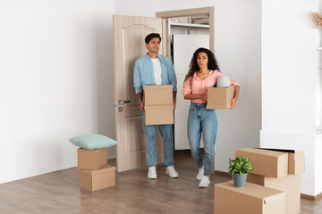 A young couple stands inside their new house, holding cardboard boxes. They appear frustrated and...