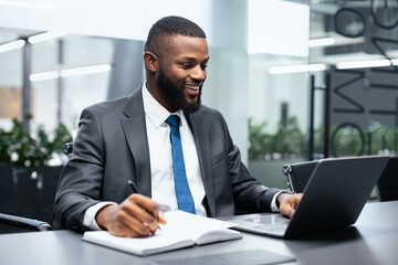 A man is seated at a desk in an office. He is focused on his laptop while taking notes in a notebook. The room has modern design and plants in the background, indicating a professional environment.