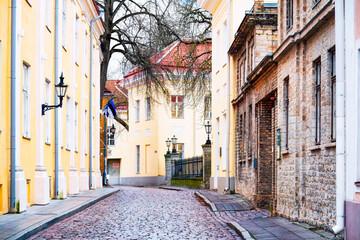 Quiet historic rhythm defines a narrow cobblestone street in the Old Town, where pastel facades, stone walls and bare winter trees shape a calm medieval city scene. Tallinn, Estonia