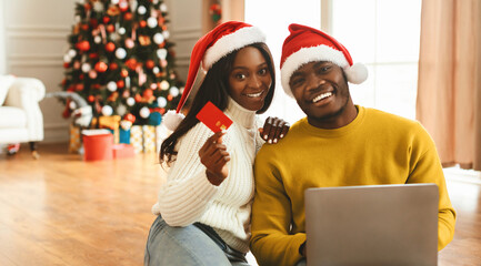A woman and a man wear red Santa hats while sitting together in a living room. They are smiling and...