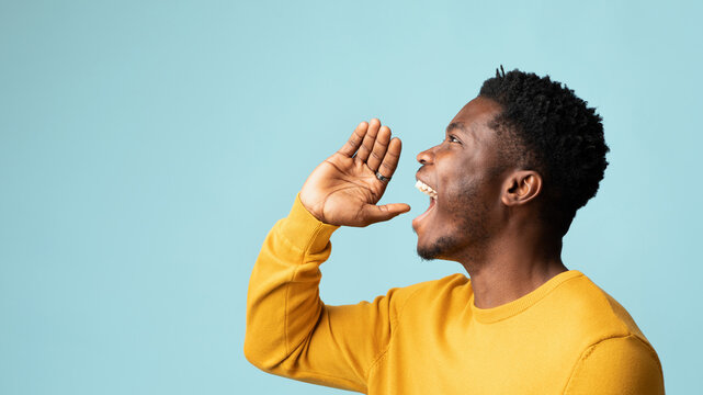 Happy african american guy posing over blue studio background, holding hand to mouth and saying something out loud, panorama with copy space. Handsome young black man making announcement, side view