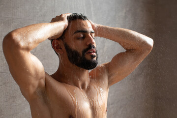 Handsome young Arabic male stands under a warm shower, applying shampoo and body wash. Water cascades over him as he experiences a rejuvenating moment in his bathroom. © Prostock-studio