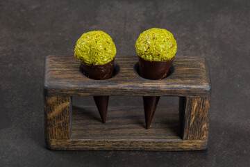 Dubai chocolate dessert. Dark chocolate cone with a ball of crispy kataifi dough with pistachio paste. On a wooden stend.  Dark gray background. Close-up.