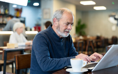 Mature man drinks coffee and works on a laptop in a cafe interior