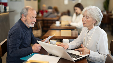 Elderly man and woman work with documents and a laptop during a business meeting in a coffee shop.