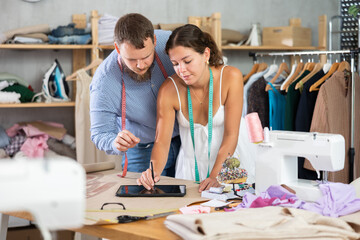 In modern sewing studio, focused young female dressmaker creating fashion design ideas on digital tablet, working closely with male colleague on new garment concepts..
