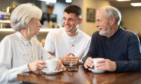 Family participants relatives sitting in cafe, talking, drinking coffee and having nice conversation. Senior couple and guy sitting at table, talking, laughing, joking, chatting