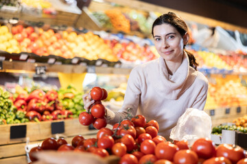 In vegetable store, young woman customer buy ripe tomato. Vegetables and fruits from different parts of world, products from local farms.