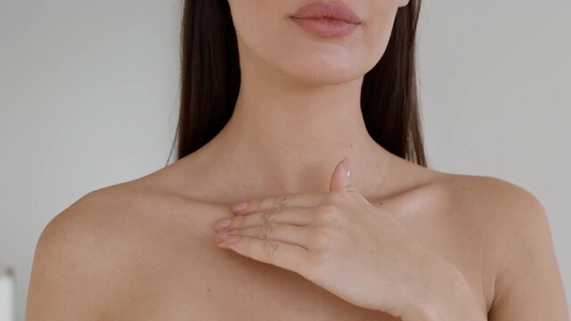 A woman stands in a bright indoor area, softly touching her collarbone with one hand. Natural light enhances the warm atmosphere, highlighting her skin and relaxed expression.
