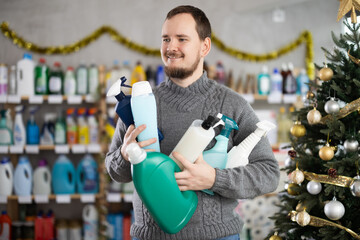 Cheerful young bearded man in cozy knitted sweater carrying various bottles of cleaning an personal...