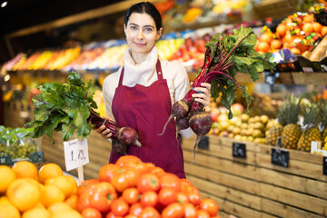 Woman staff with beetroot in hands, vendor demonstrate, adds and displays goods. In sales area of vegetable store, seller restructures display case.