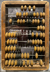 Close-up of a vintage wooden abacus with multiple rows of round beads in natural and dark tones, mounted on metal rods within a rectangular wooden frame, displayed vertically.