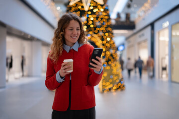 Happy woman checking smartphone holding coffee in mall