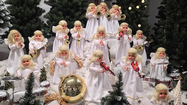 A festive display of numerous white-clad angel figurines playing musical instruments, set among decorated Christmas trees and holiday ornaments in a cozy winter scene. Video 4k