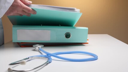Doctor's hand selecting files from a binder on a white desk, with a blue stethoscope lying next to the organized medical paperwork, symbolizing healthcare administration and patient data management