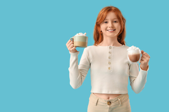 Teenage redhead girl with cups of sweet cocoa on blue background