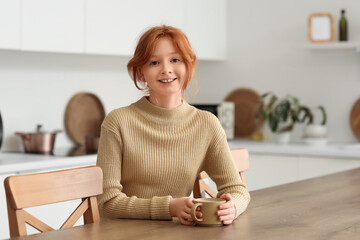 Teenage redhead girl with cup of sweet cocoa sitting at table in kitchen
