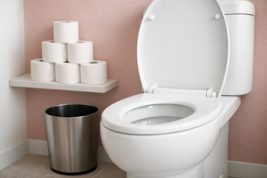 Clean white toilet with open lid, stainless steel trash bin and stacked toilet paper rolls against soft pink bathroom wall