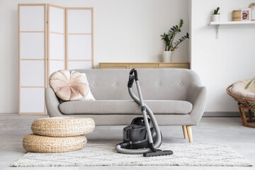 Modern vacuum cleaner with poufs on carpet near sofa in living room interior