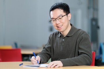 A male Chinese student concentrates while writing with a blue pen on paper at a desk. He appears focused in taking an exam, quiz, or completing homework in a contemporary educational setting.