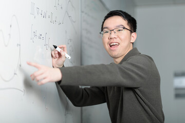 A Chinese college student smiles enthusiastically while explaining math equations on a whiteboard with a marker. He is teaching or presenting the solution to a math problem in the classroom.