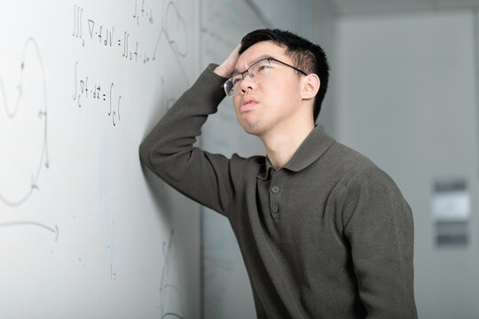 A Chinese college student scratches his head in confusion while looking at math equations on a whiteboard. He is stuck and frustrated while trying to solve a challenging math problem in the classroom. - Powered by Adobe