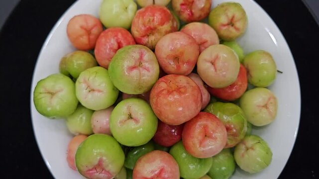 Top-down shot of fresh red and green acerola cherries on a white ceramic plate.