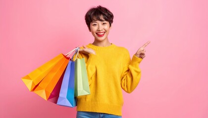 Portrait of cheerful young asian woman holding colorful shopping bags and pointing finger at copy space on pink background. Retail concept
