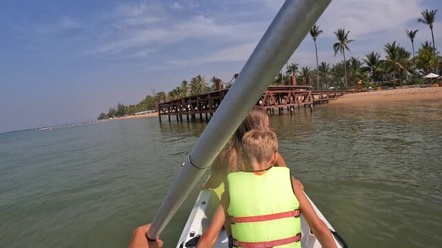 First-person view - a hand with an oar, a kayak goes through calm water to a sandy beach with palm trees. Family vacation, summer, resort.
