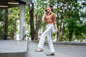 Obraz premium Full body shot of asian woman student talking on phone carrying laptop and walking on stairs in park