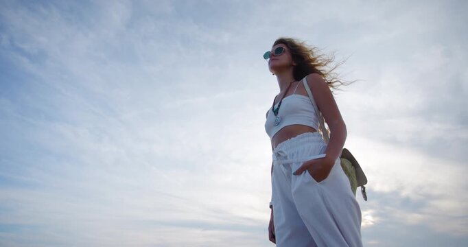 Portrait of a woman enjoying a warm day at the beach, dressed in stylish white attire with sunglasses and a backpack by her side under a cloudy sky
