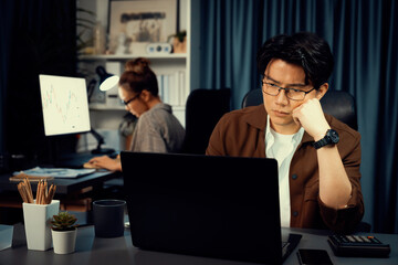Serious face of thinking businessman focusing laptop to report data analysis social media online project with coworker analyzing stock at back side at over late night time at modern office. Infobahn.