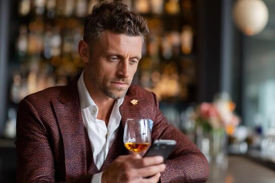 Man at the Bar: A sophisticated man, impeccably dressed, sits at a bar, a glass of amber liquid in hand, engrossed in his phone. The ambiance suggests a moment of contemplation.