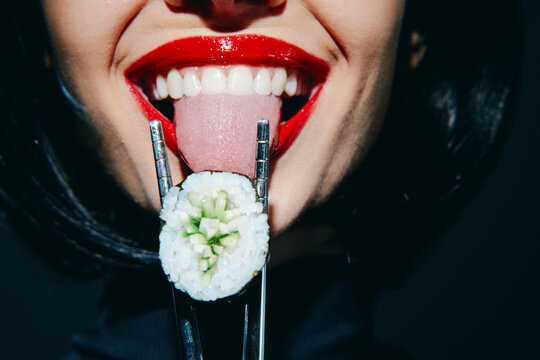 Seductive woman with red lips teasingly holding sushi with chopsticks, showcasing playful emotions against a dark background.