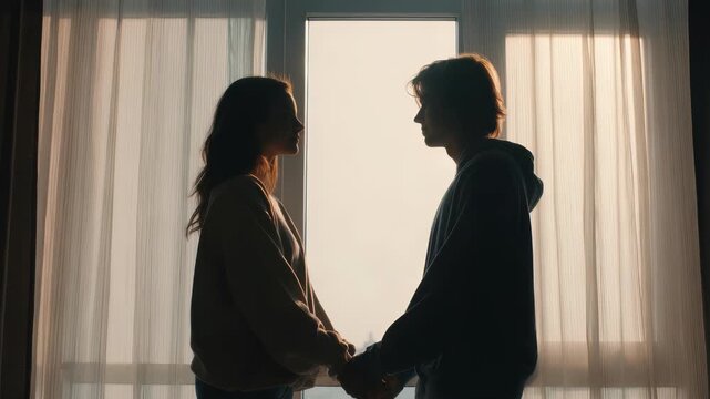 A young man and woman stand close, holding hands and gazing at each other in silhouette against a bright window. The scene conveys intimacy, love, and a serene, peaceful mood