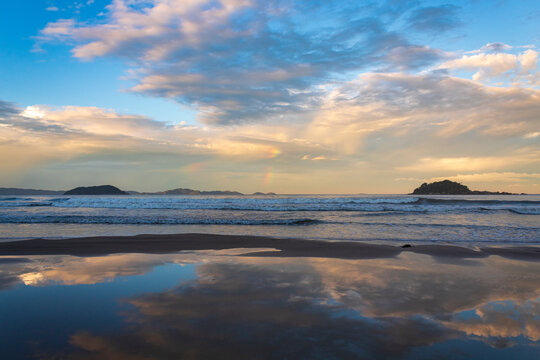 reflexo do c&eacute;u e o p&ocirc;r-do-sol na Praia Palmas do Arvoredo cidade de Governador Celso Ramos Santa Catarina Brasil regi&atilde;o da Grande Florianopolis  ao fundo um arco-&iacute;ris na costa de Bombinhas  Mariscal  