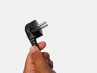 Close-up image of a human hand holding a black European two-pin power plug, isolated on white background, representing household electricity and electrical connection.