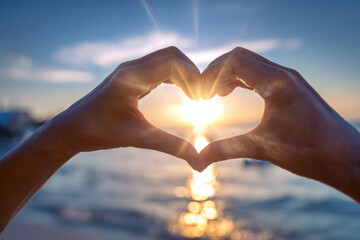  beautiful hands forming a heart shape at dramatic soft blurred sunset with sunrays by the sea, a symbol of love and romance for Valentine background.