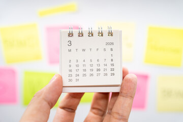 Hand holding MARCH 2026 mini calendar with blurred colorful sticky notes for planning concept. project tracking, monthly goal setting, and time management in a creative workspace.