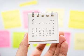 Hand holding February 2026 mini calendar with blurred colorful sticky notes for planning concept. project tracking, monthly goal setting, and time management in a creative workspace.
