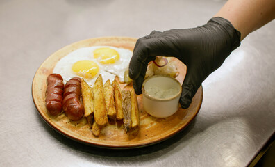 Serving lunch. Meal plate. Hand in black gloves putting ingredients for hearty breakfast. Chef preparing fried potato egg sausage vegetable sauce bread.
